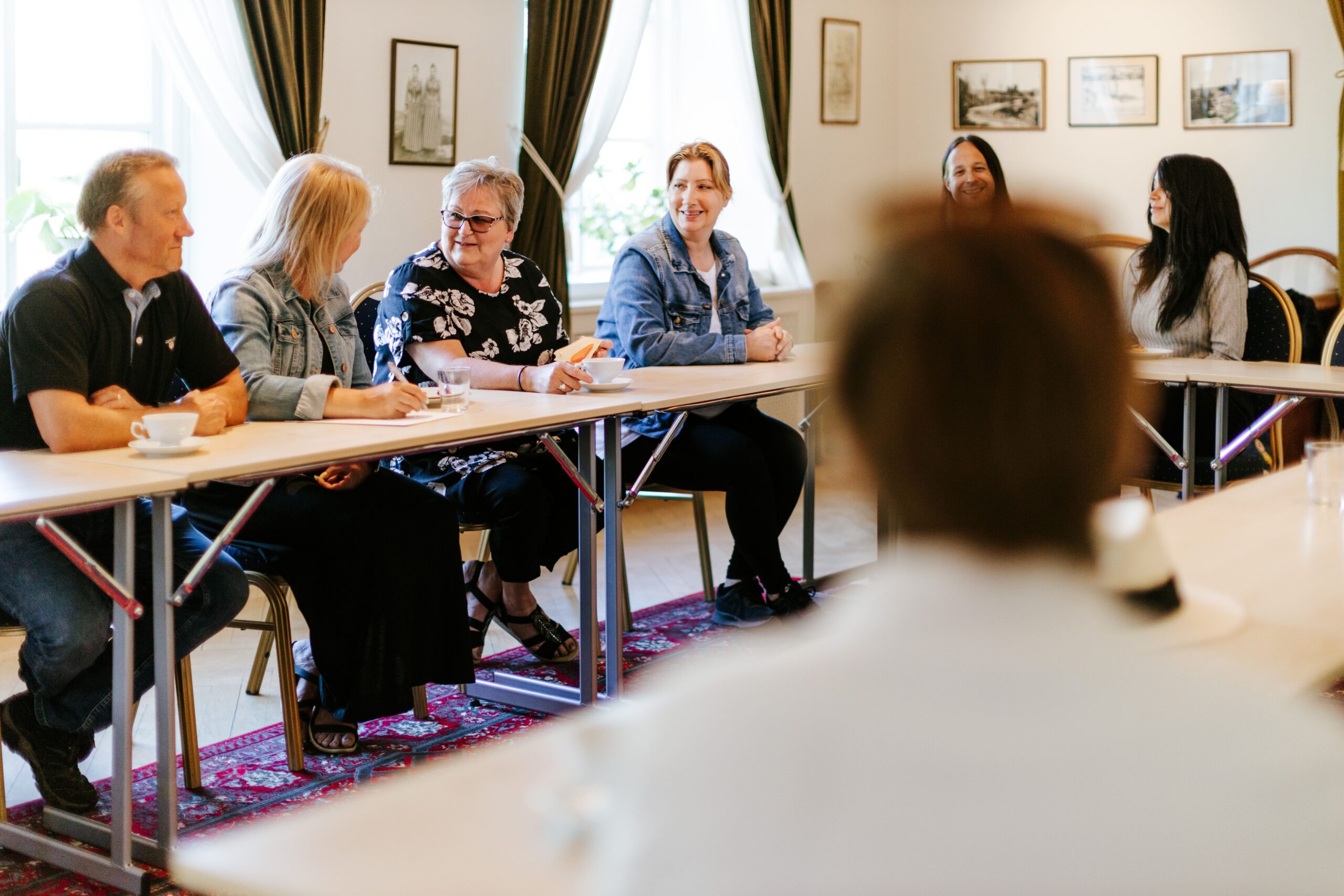 Deltagare i en konferensdiskussion, sittande vid bord, med fikabröd och en ljus, inbjudande lokal.