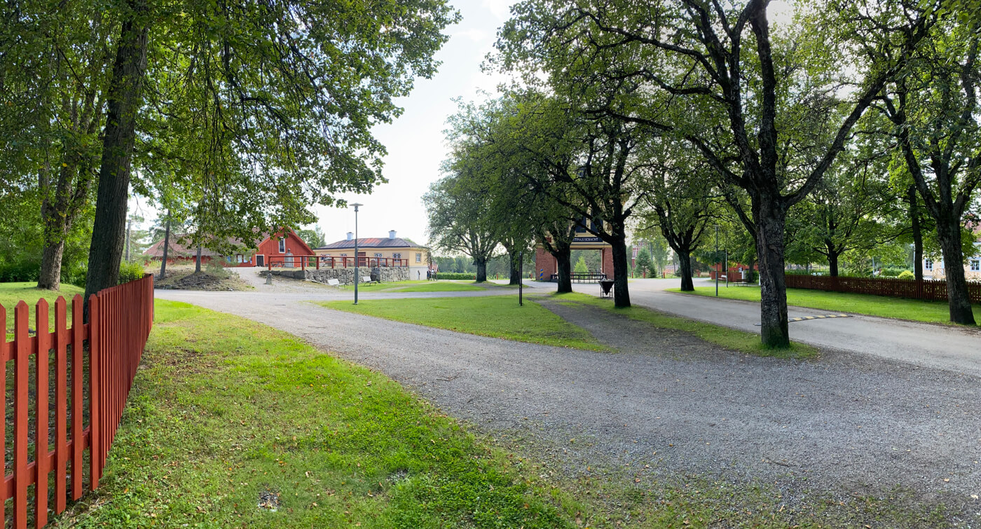 Grön park med träd och röd staket, som leder till en byggnad vid Sala Silvergruva. Lugnt landskap i dagligt ljus.