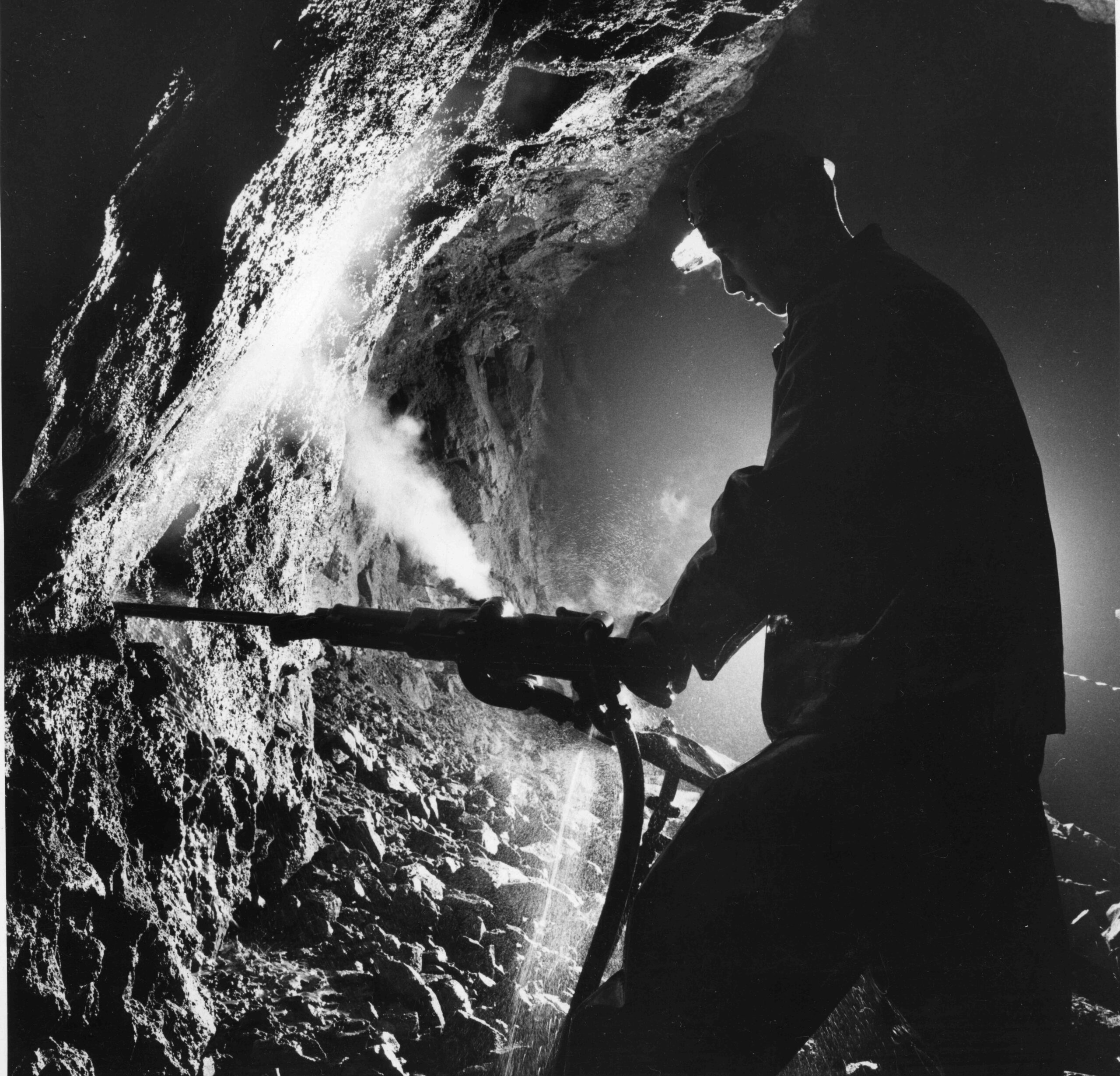 Gruvarbetare som arbetar med en borrmaskin i en mörk tunnel, omgiven av stenar och ånga i Sala Silvergruva.
