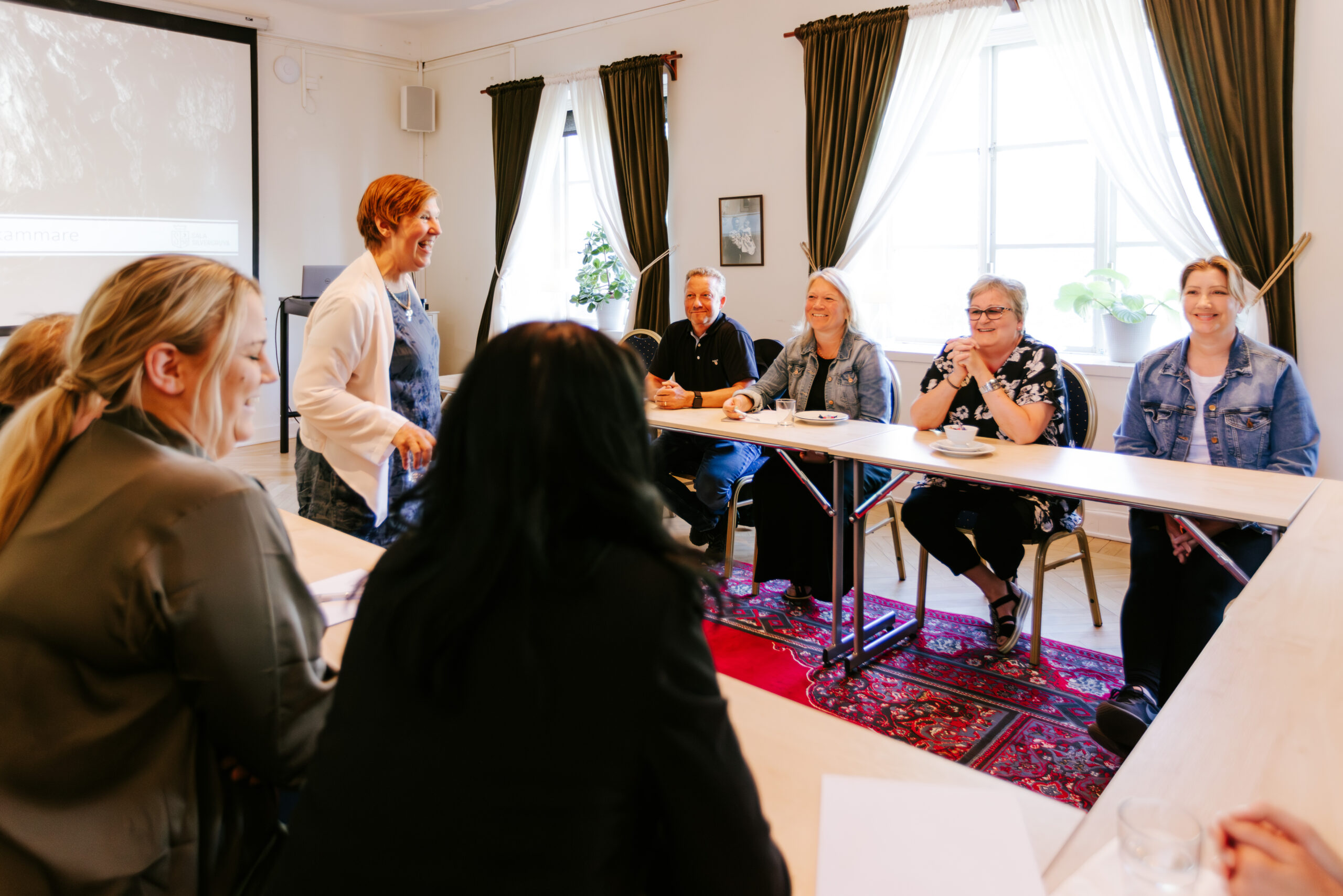 Konferenssituation med deltagare vid ett bord, ledd av en kvinna, i en ljus och inbjudande lokal.