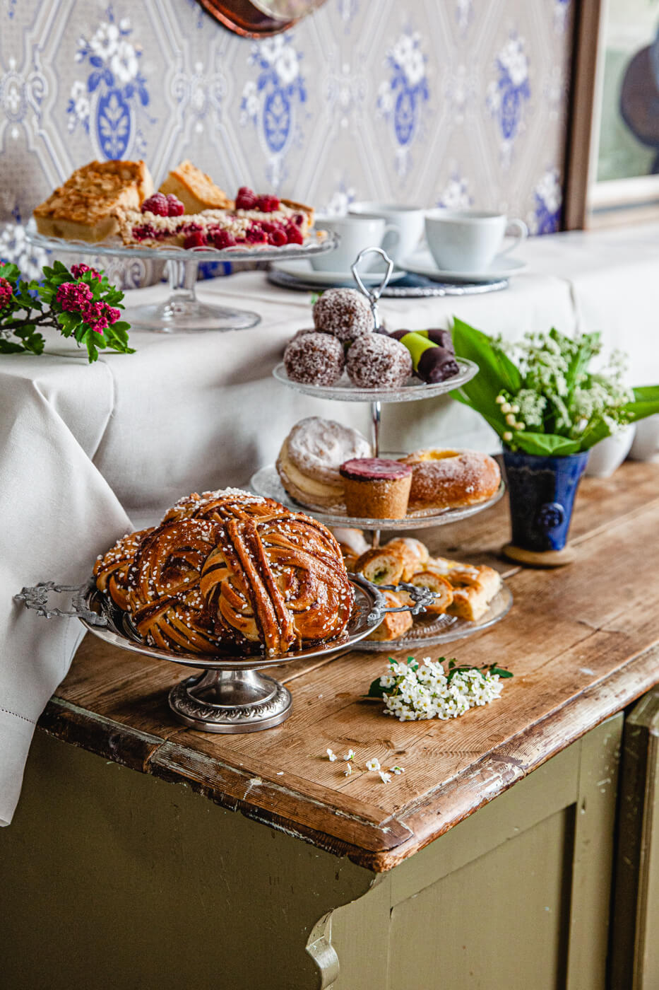 Ett dignande fikabord med bakverk som bullar, småkakor och tårtor, omgiven av blommor i en inbjudande miljö.