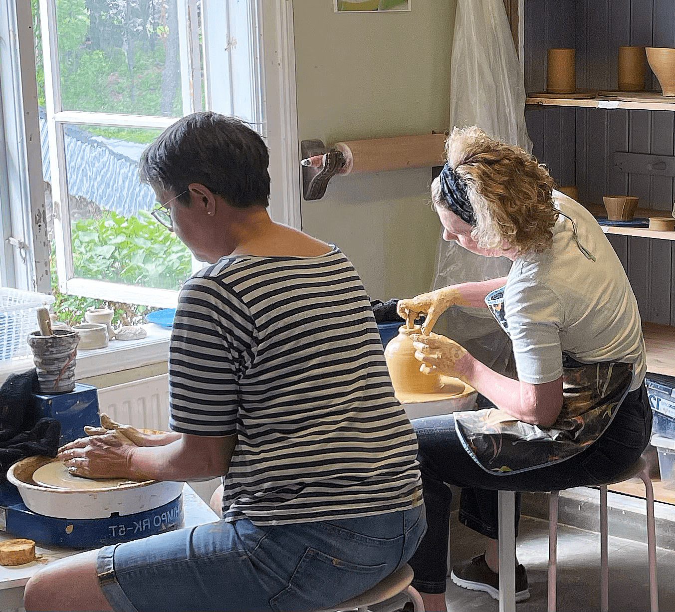Två kvinnor sitter vid en verkstad och drejar keramik, omgivna av olika skapelser och ljusinsläpp från ett fönster.