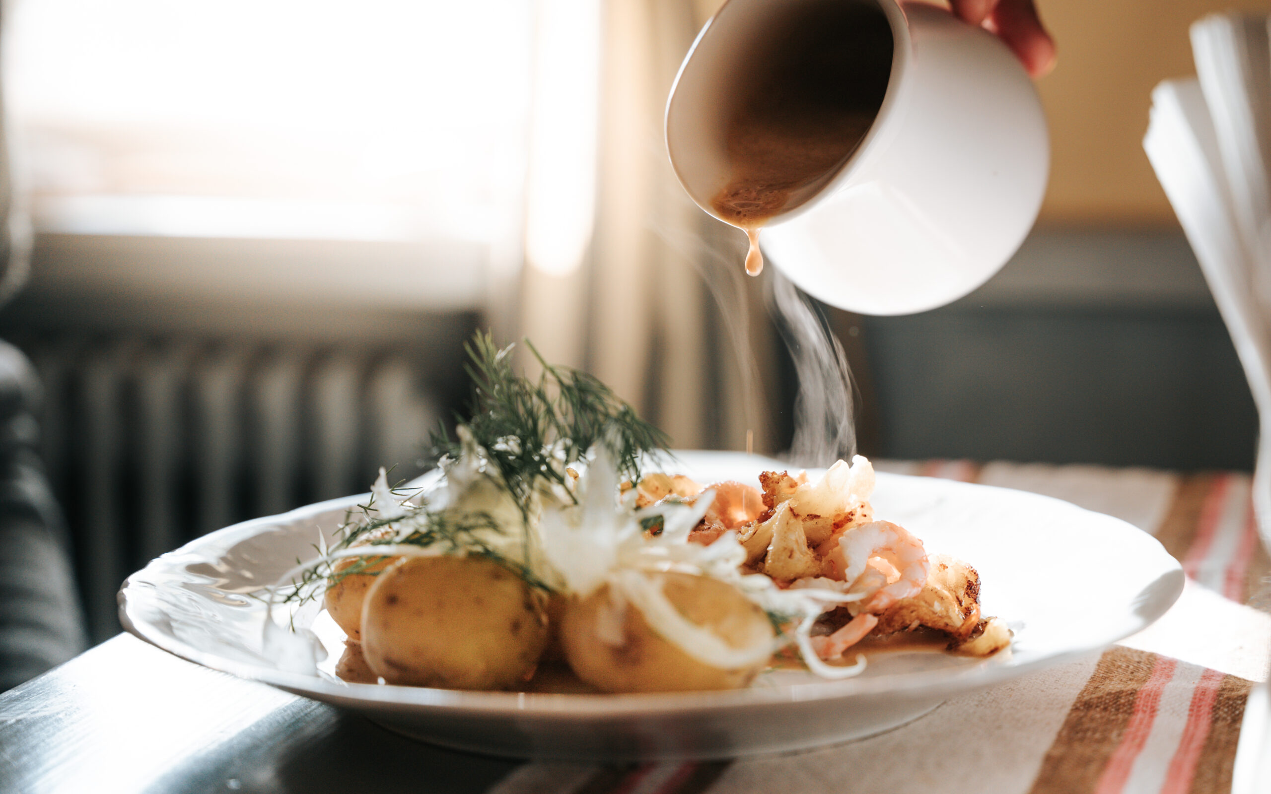 En hand häller sås över en tallrik med potatis, lax och grönsaker i en ljus och hemtrevlig miljö.