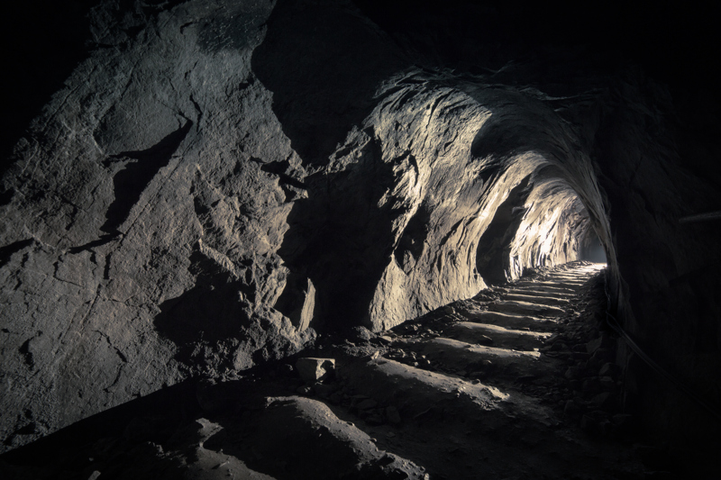 Dimly lit mine tunnel with rocky walls and a path illuminated by light at the end, showcasing the underground environment.